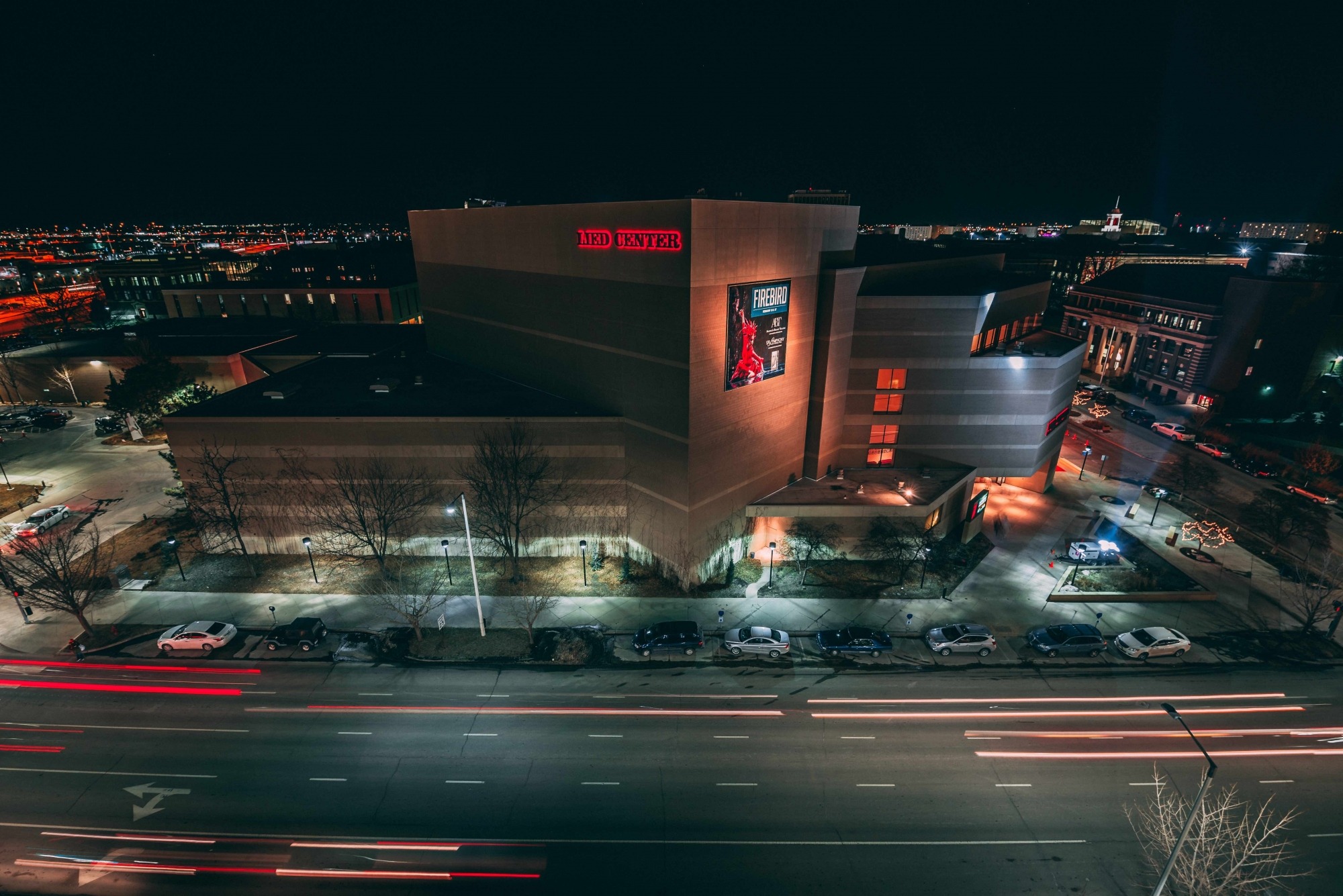 Image of the Lied Center building at night taken from the roof of a nearby building showing searchlights pointed towards the sky and car lights streaking across the road